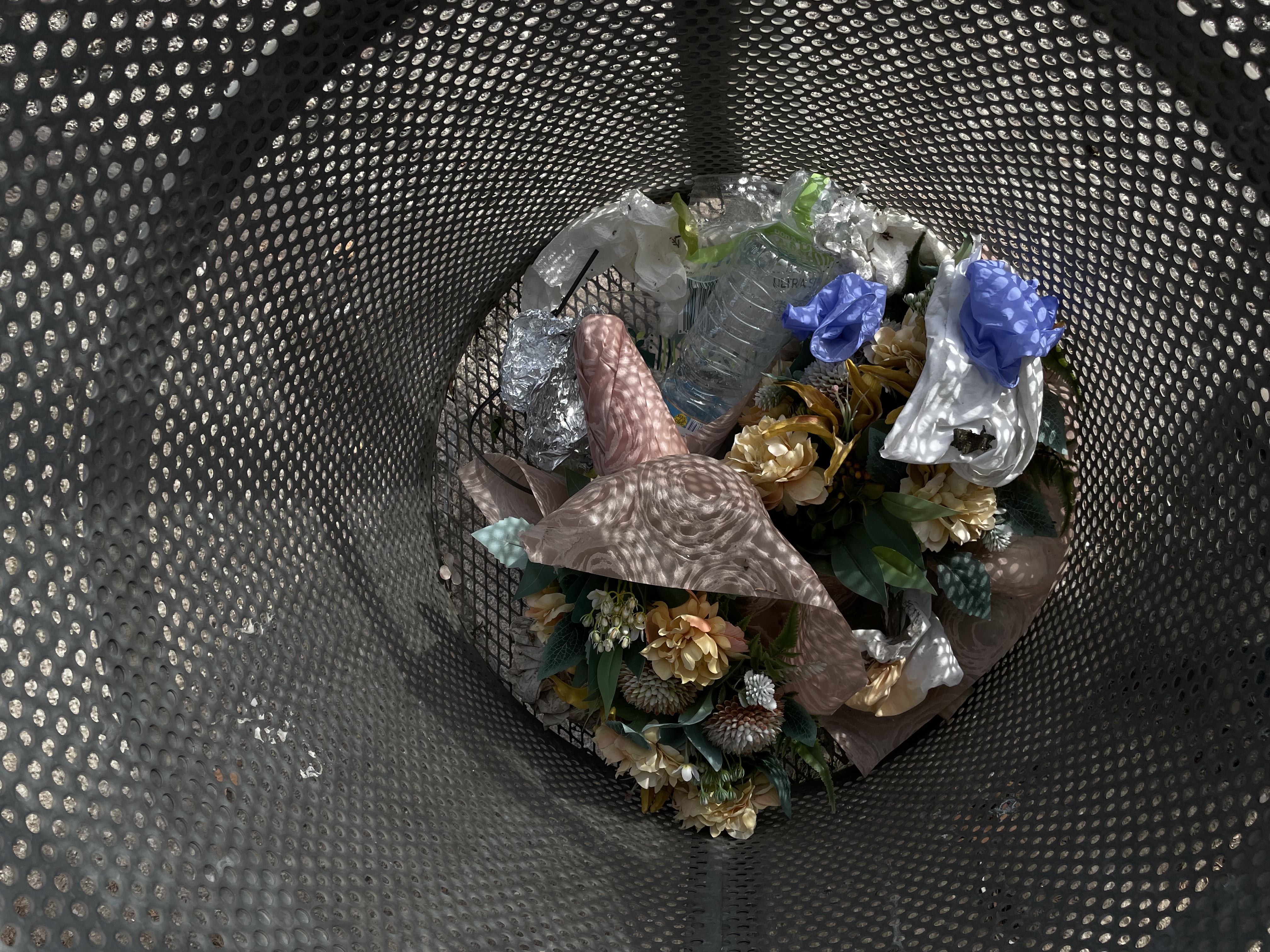 view into a metal trash can filled with plastic flowers, plastic bottles, tissues, and other trash. The trash can is made of perforated metal, through the holes light shines in a dotted pattern onto the trash.