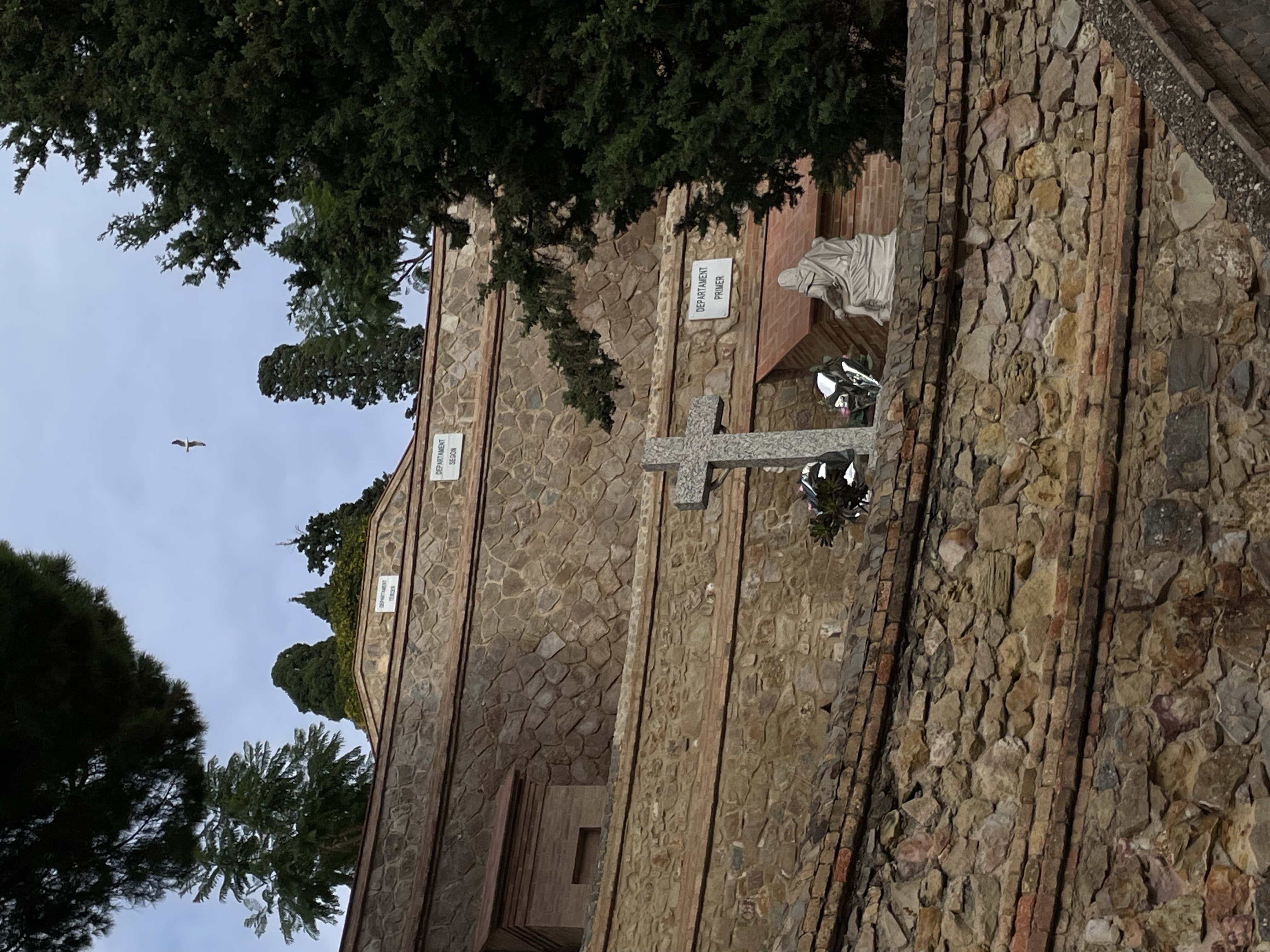 looking up a hill, four stone walls, layered behind each other. the three in the back have plaques reading: department primer, department segon, department terer. behind, above the first wall, before the second wall, is a cross with flowers and a small statue, and a tree to the right